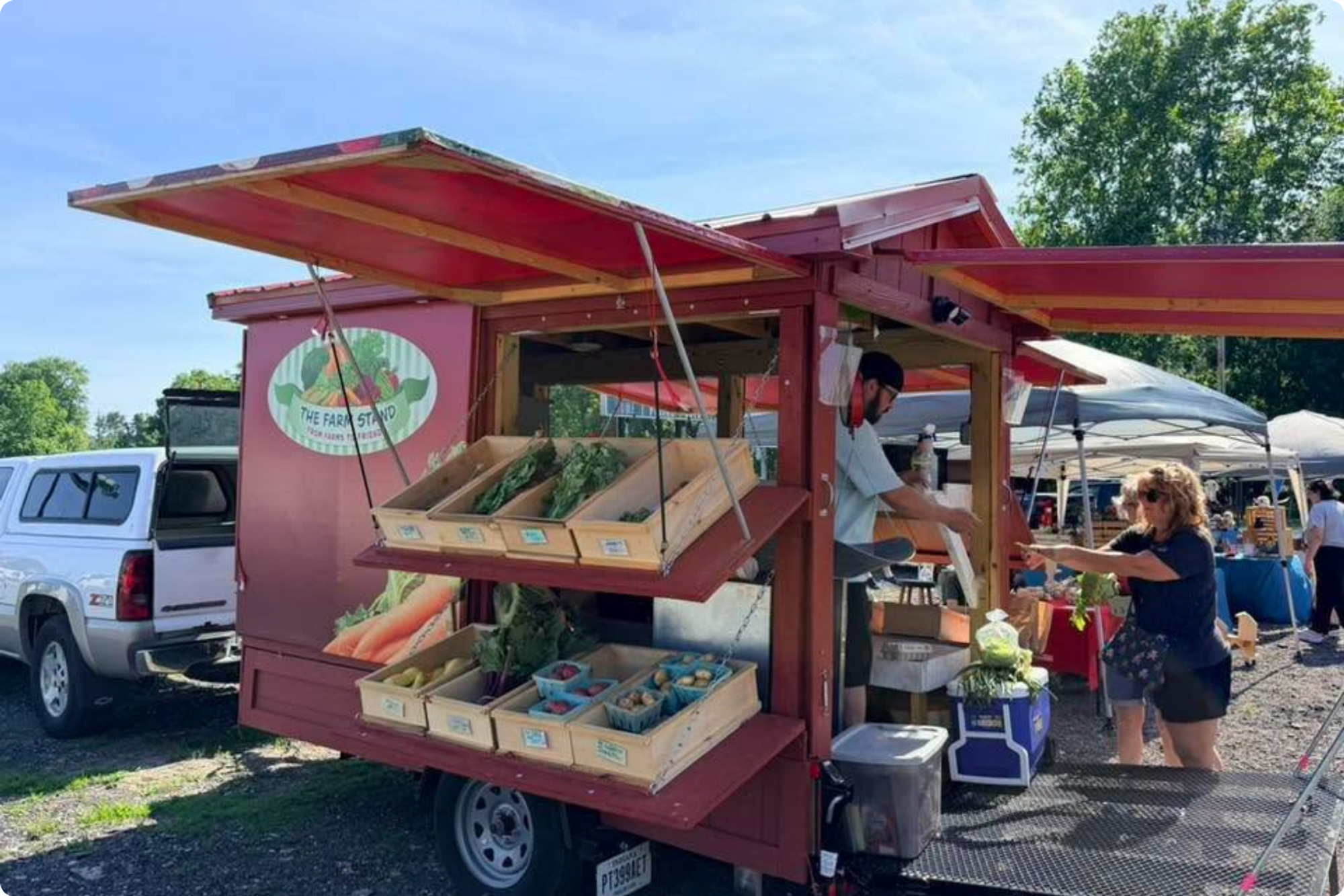 Women purchase items from the Farm Stand, which is set up with pop-out shelves and awnings for shade.
