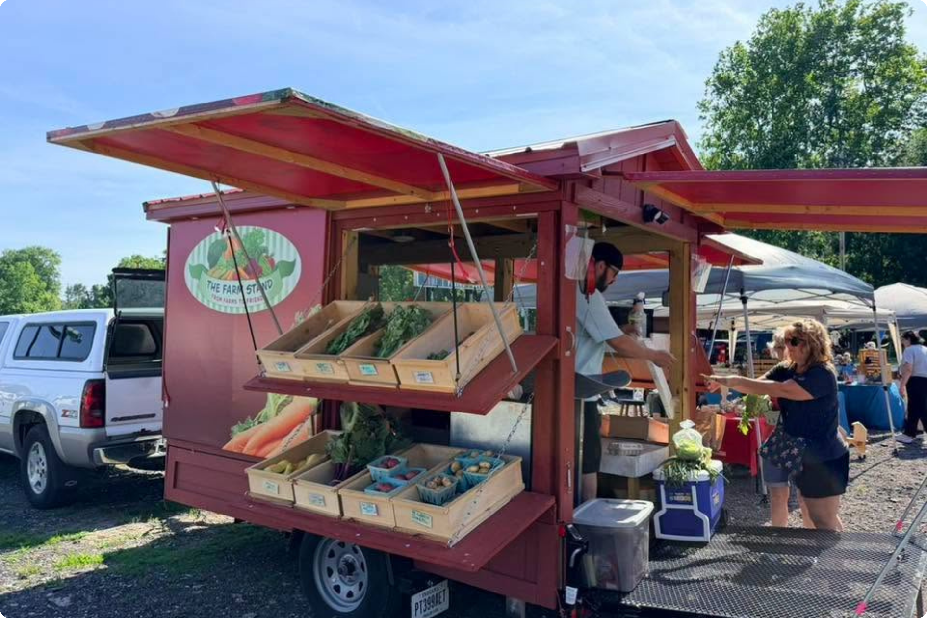 Women purchase items from the Farm Stand, which is set up with pop-out shelves and awnings for shade.