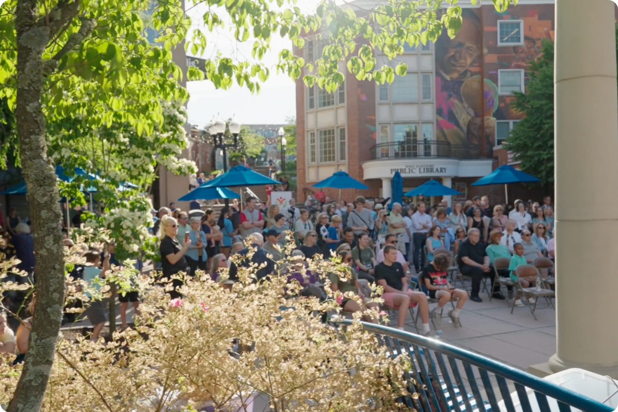 A crowd gathers for the unveiling ceremony of the Voices in Color mural, shot through trees in the foreground