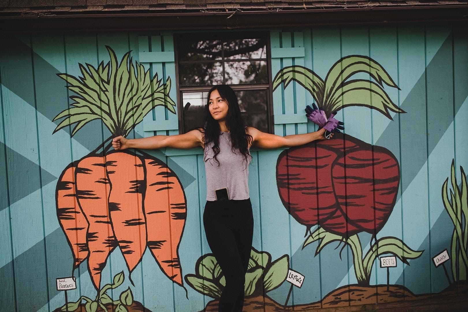 A woman stands in front of a mural in the Community Rocks Vegetable Garden of turnips and carrots and stands in front to look as though holding the vegetables.