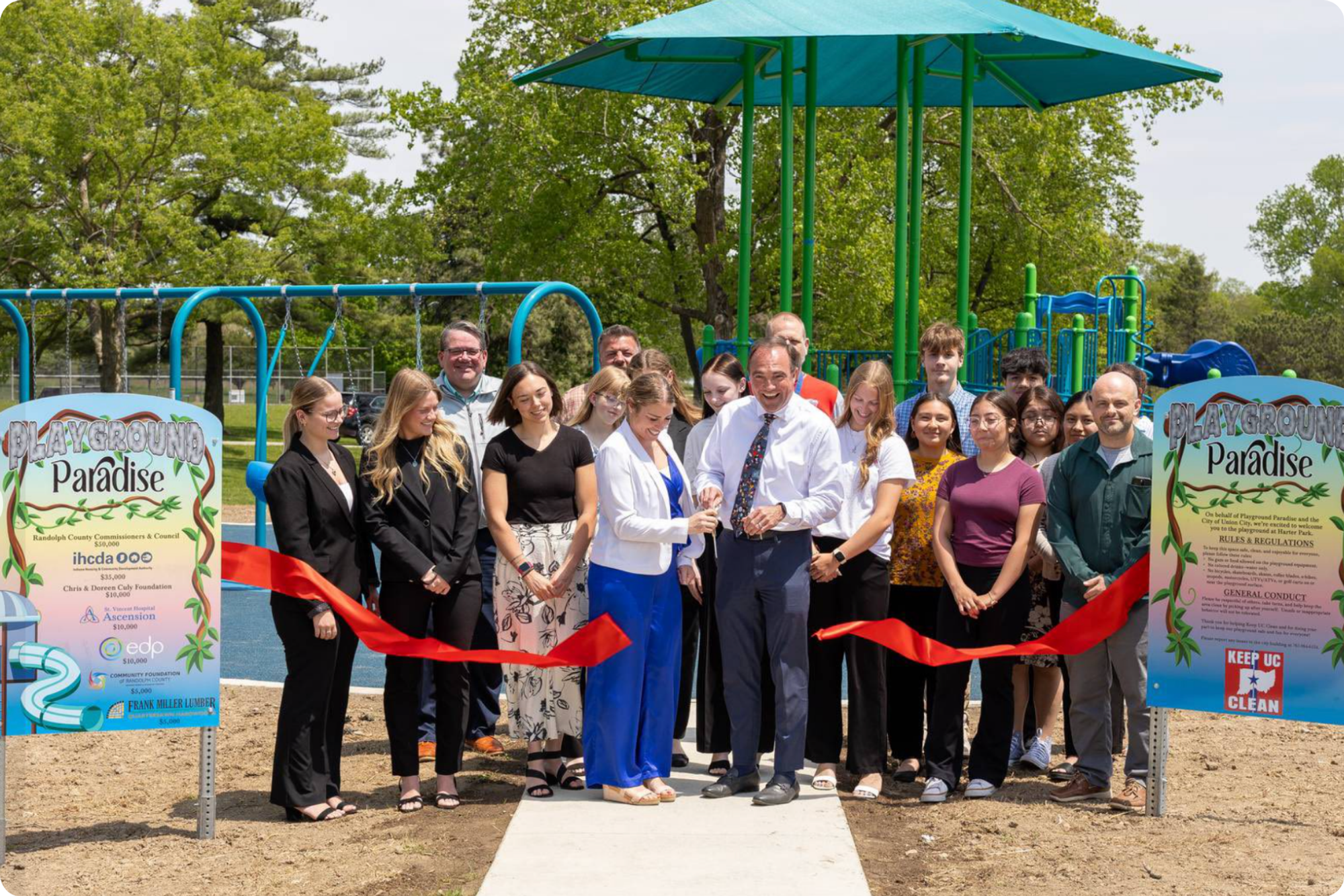 Teens and adults gather in front of the new playground between two signs and behind a snipped red ribbon.