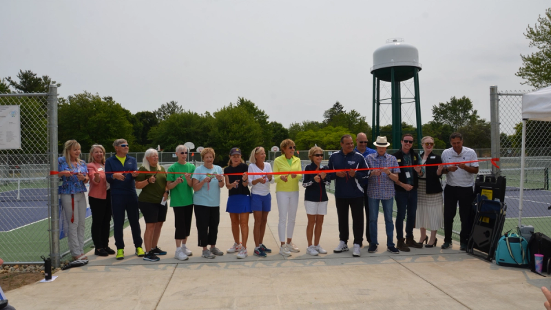 Sixteen people from Sue’s crowdfunding team stand in behind a red ribbon spanning the opening of a large pickleball court