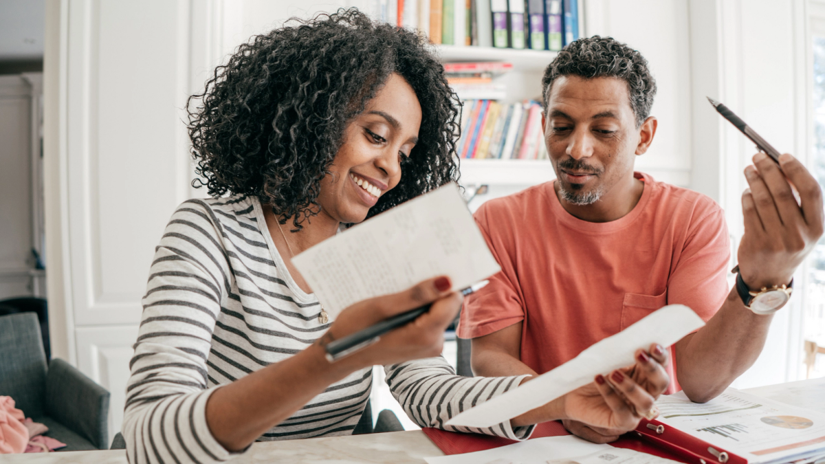 A middle-aged Black woman in a striped shirt reviews receipts with a Black man in a salmon t-shirt.