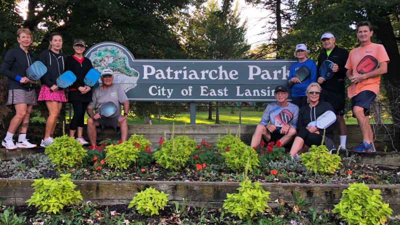 Nine people in athletic clothing posing with pickleball paddles in front of the sign for Patriarche Park in East Lansing.