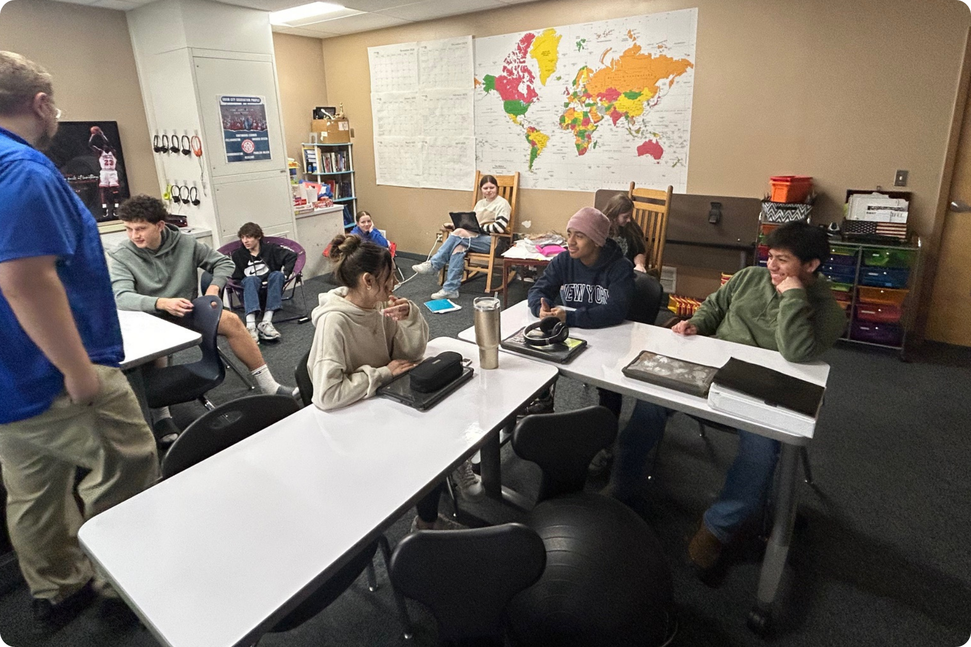 A collection of students are clustered around classroom tables to work in small groups under teacher supervision