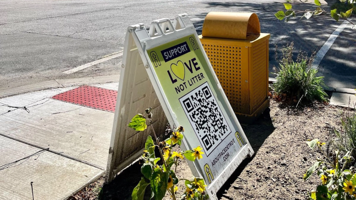 An A-Frame sign reads, “Support Love Not Litter,” next to a yellow trash can in East Village, Detroit. 