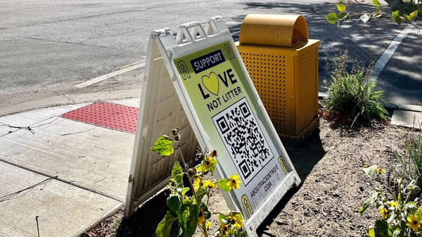 An A-Frame sign reads, “Support Love Not Litter,” next to a yellow trash can in East Village, Detroit.