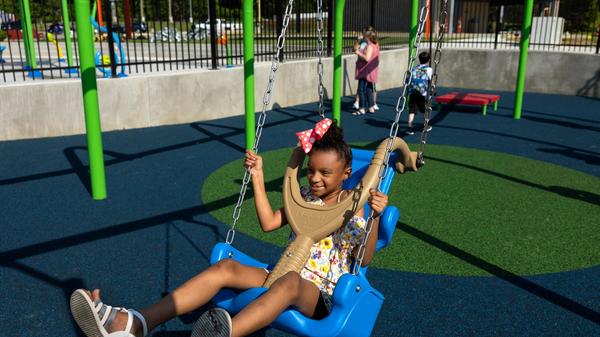 A young Black girl with white sandals and a pink bow in her hair swings on the accessible playground at Kevin Hammersmith Memorial Park. 