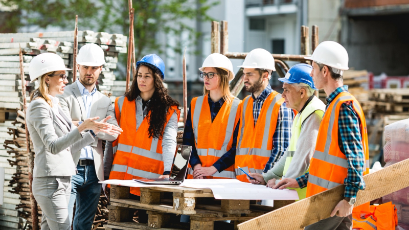A group of people stand around building plans in hard hats and safety vests. 