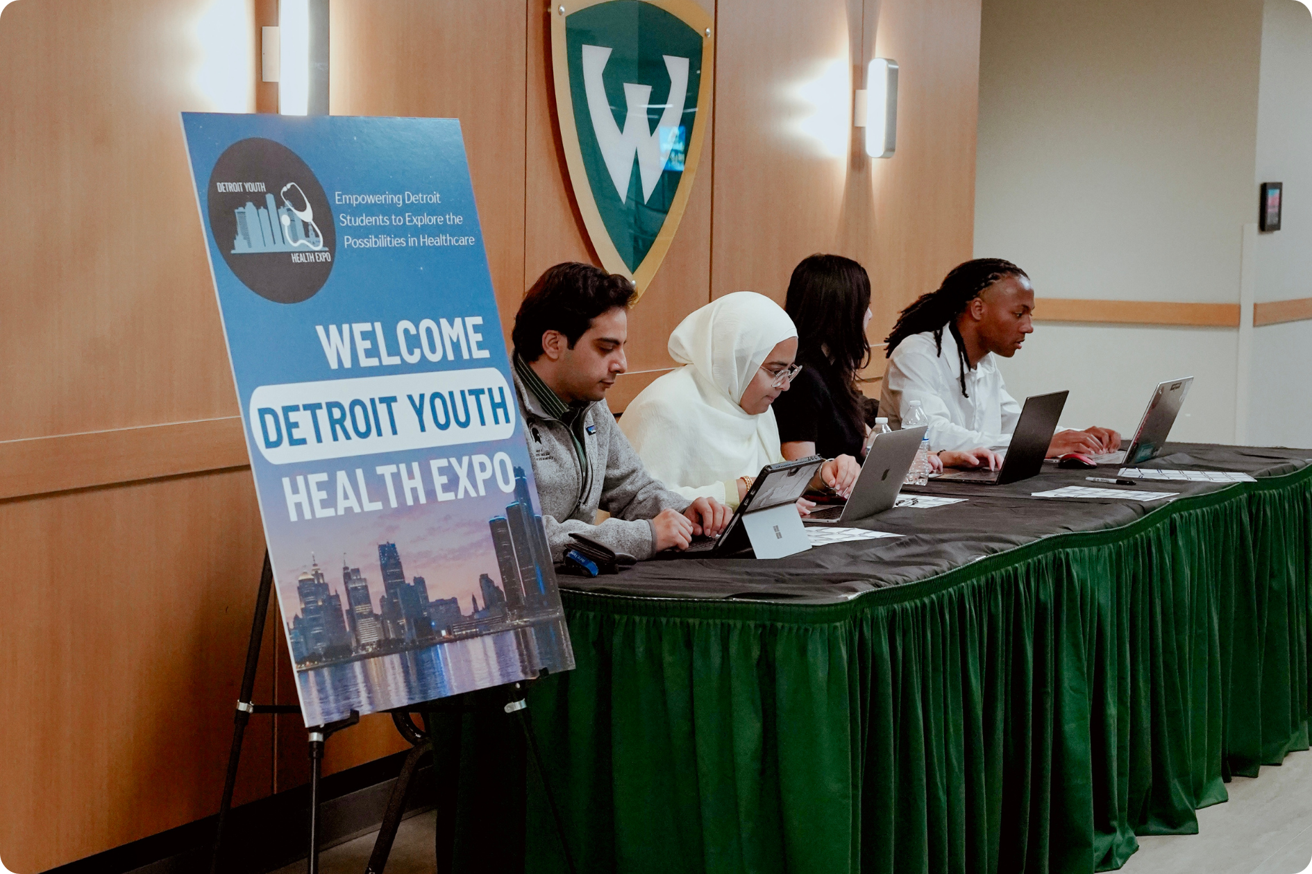 Four university students sit with tablets and laptops at a long table, beside a large sign reading "Welcome, Detroit Youth Health Expo"