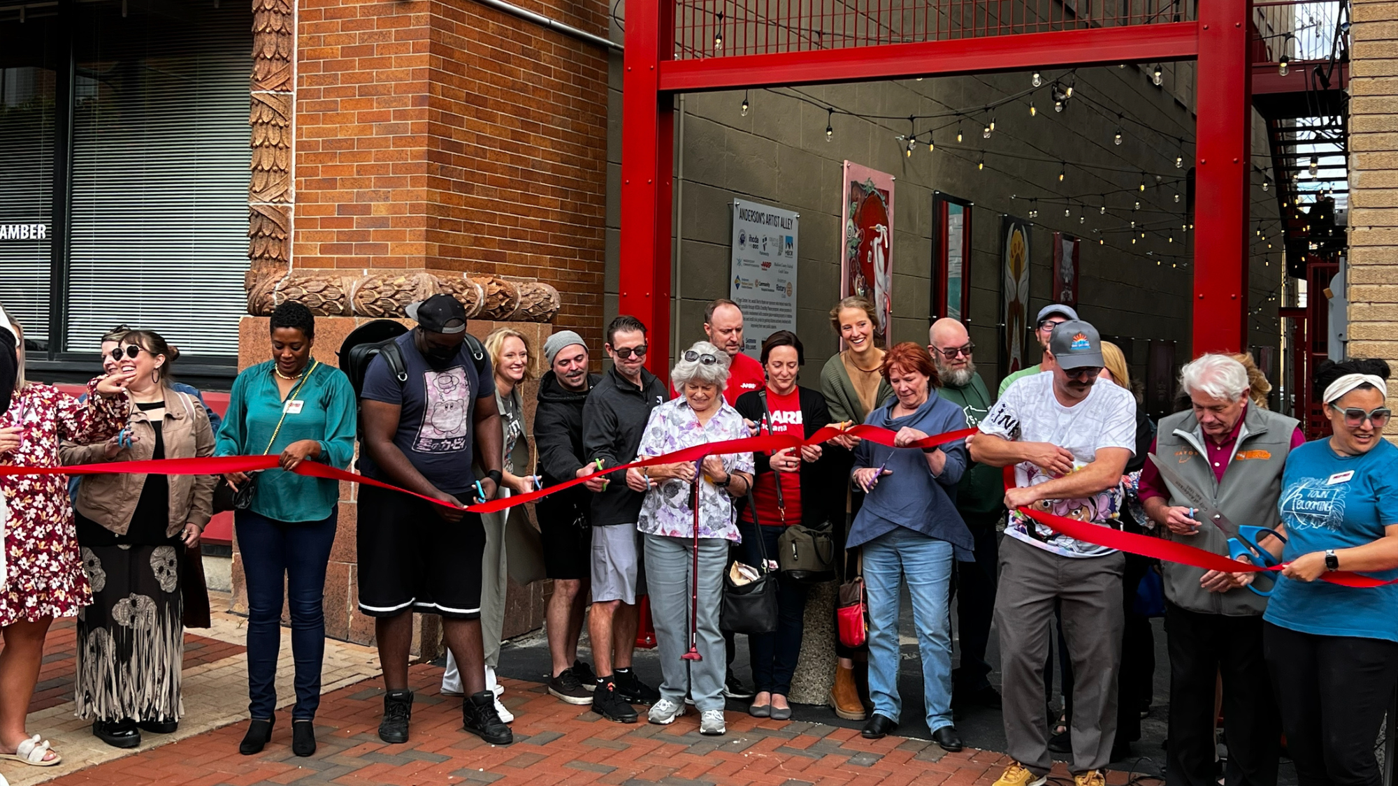 Community members stand in a long line across the archway to the artist alley holding a bright red ribbon. 