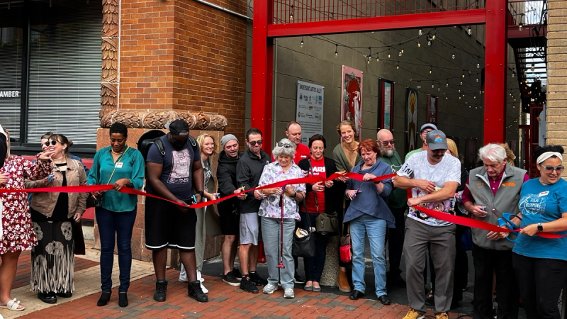 Community members stand in a long line across the archway to the artist alley holding a bright red ribbon. 