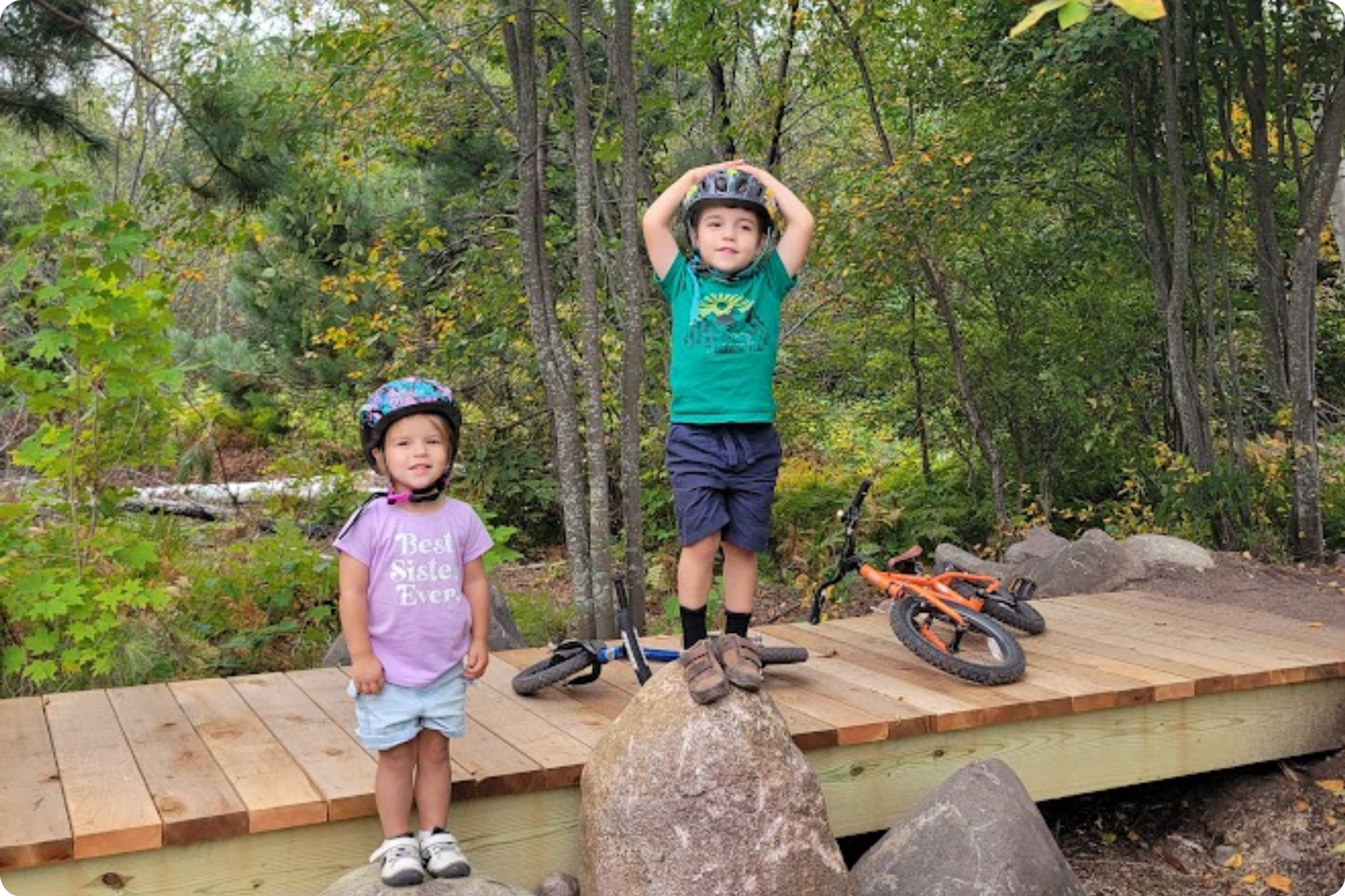 Two children pose on a boardwalk with their bicycles behind them.