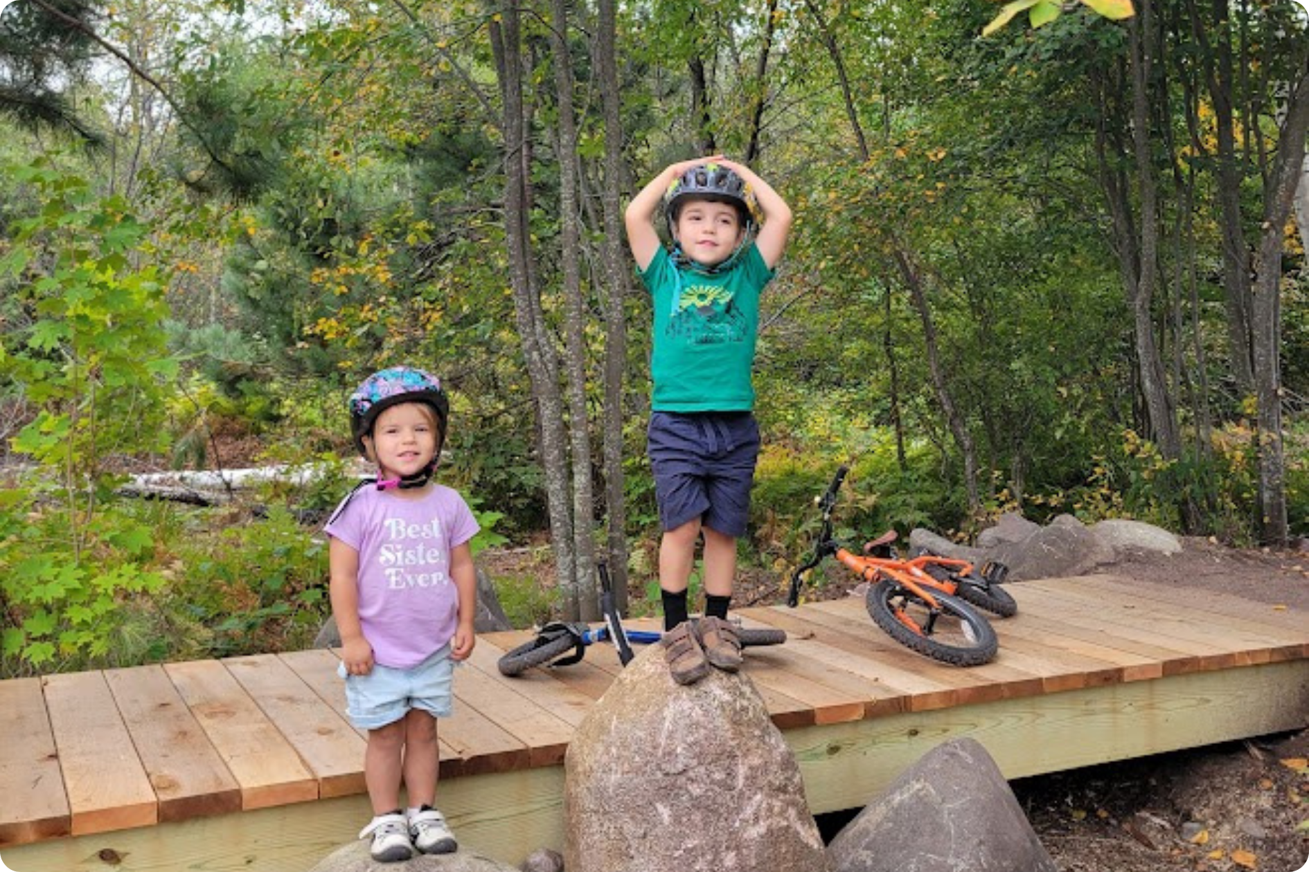 Two children pose on a boardwalk with their bicycles behind them.