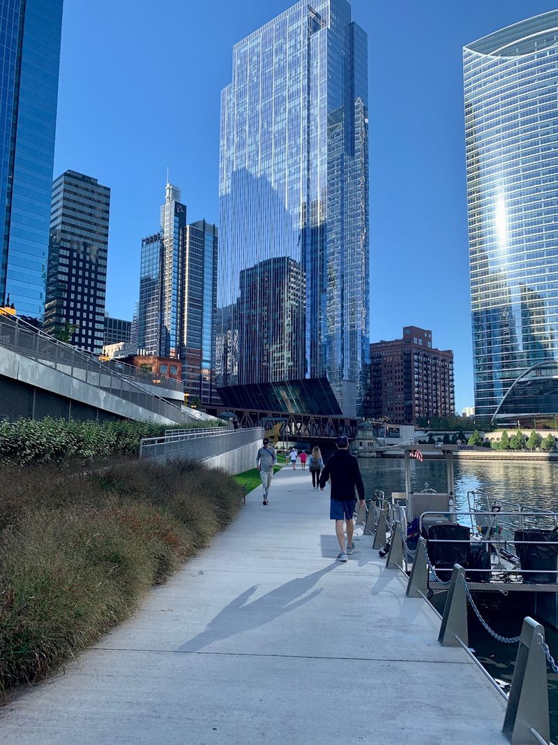 Pedestrians walk along the river beneath the city skyline with towering buildings.