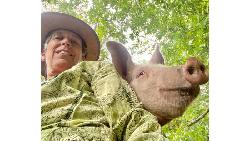 Rebecca Miller, wearing a green blouse and a wide brim hat, takes a selfie with her happy pink pig. 