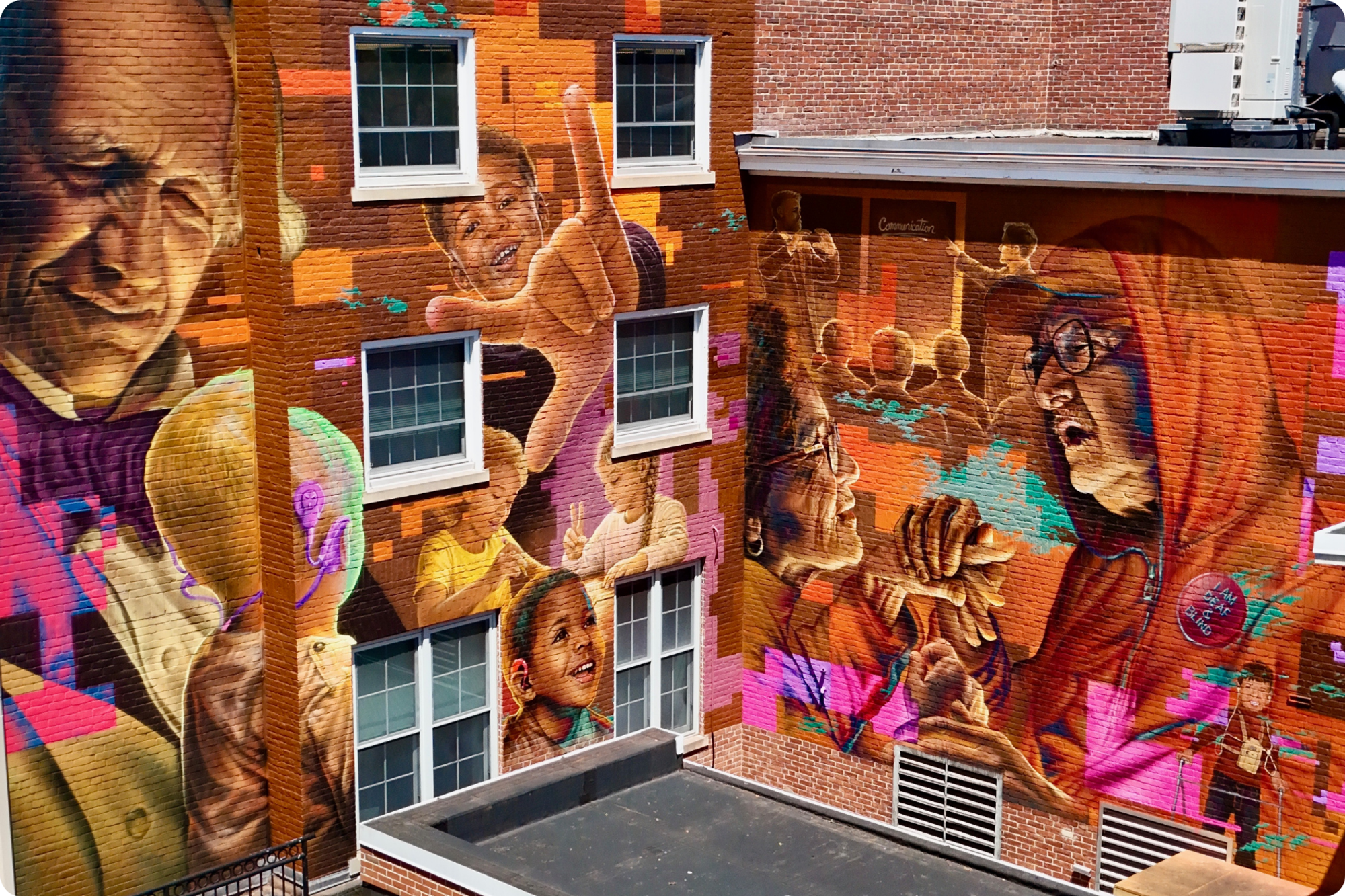 The final result of the Voices in Color mural: a vibrant display representing elements of the DHH community, with portraits of Deaf people of all ages decorating the brick building.