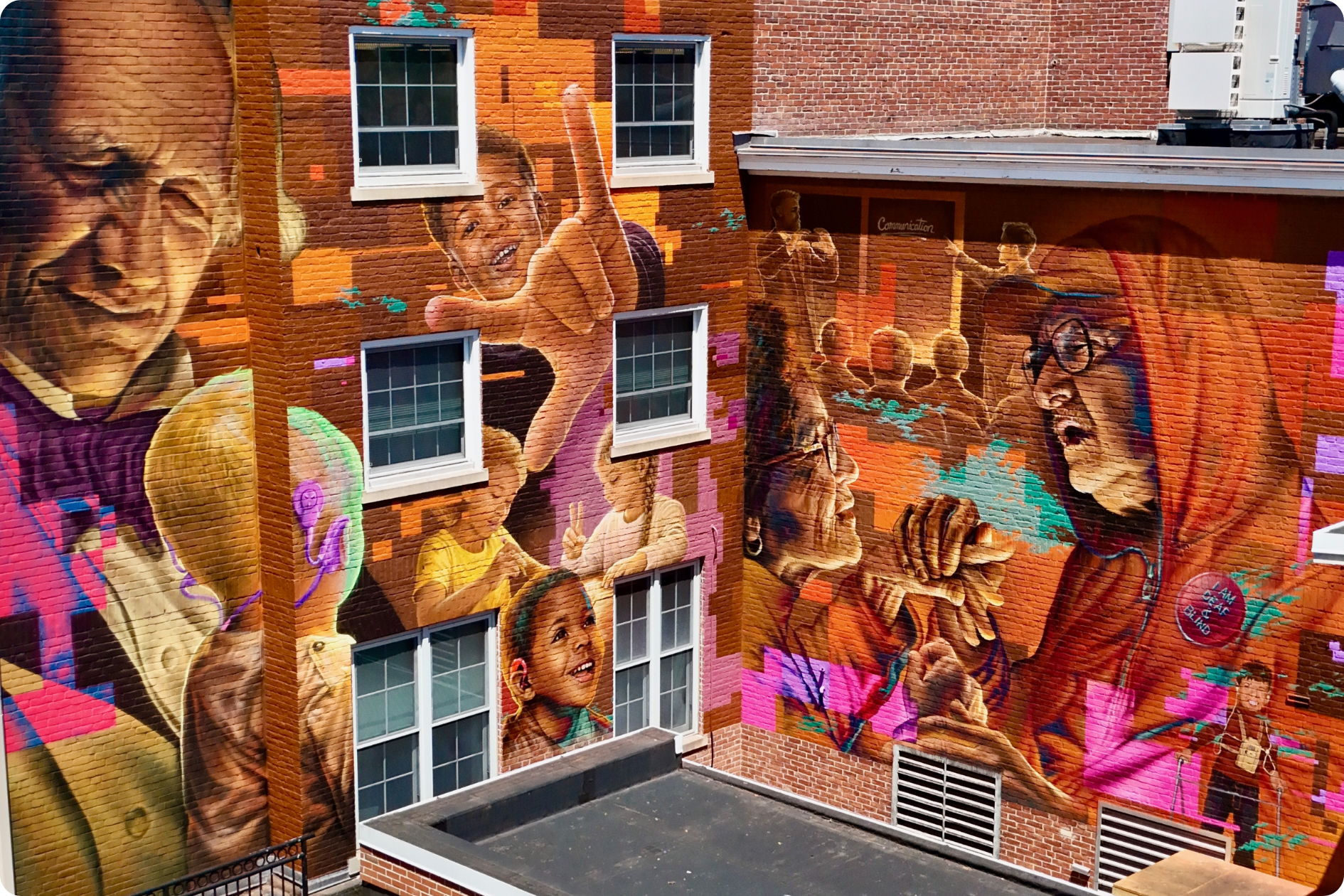 The final result of the Voices in Color mural: a vibrant display representing elements of the DHH community, with portraits of Deaf people of all ages decorating the brick building.