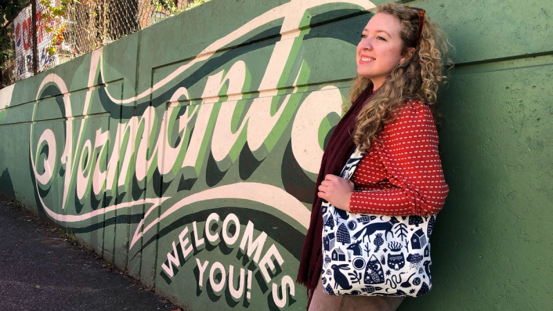Camryn Greer, Director of Programs & Impact, stands in front of the High Street mural that reads "Vermont welcomes you!"