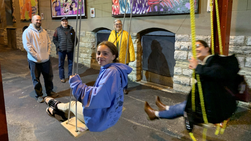 Five people in an alley lined with framed artwork and lights. Three people stand and look at the camera, while two people sit on swings facing away from the camera and look back.