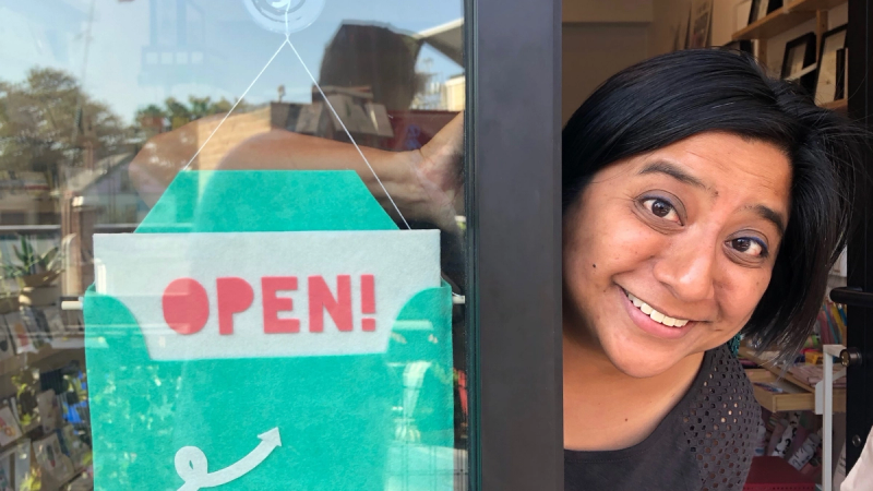A woman of color with black hair smiles next to her open business sign. 