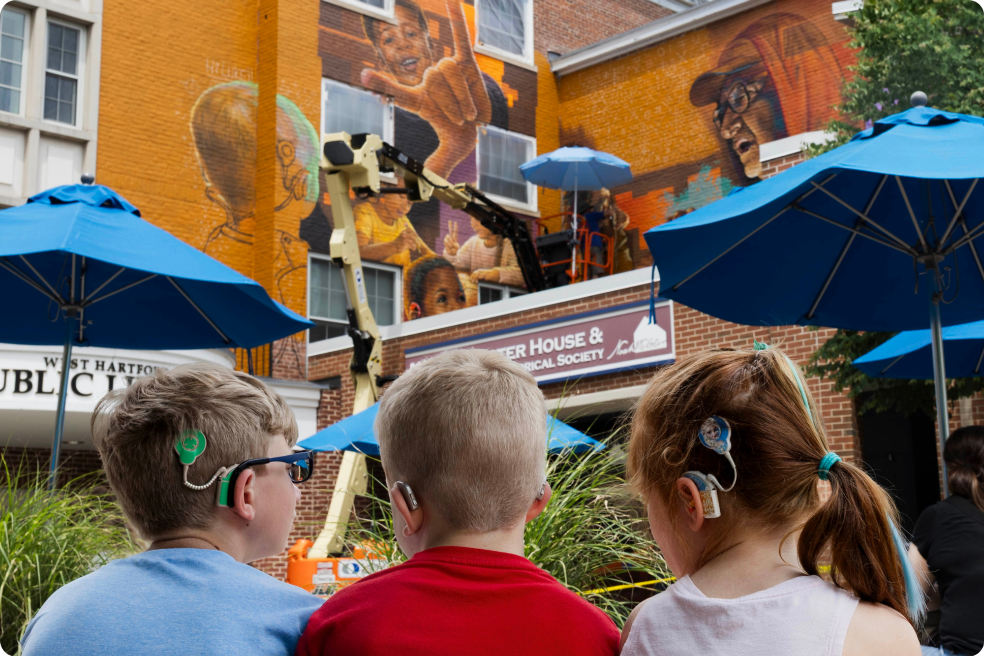 Three children with cochlear implants and hearing aids look toward the mural in progress