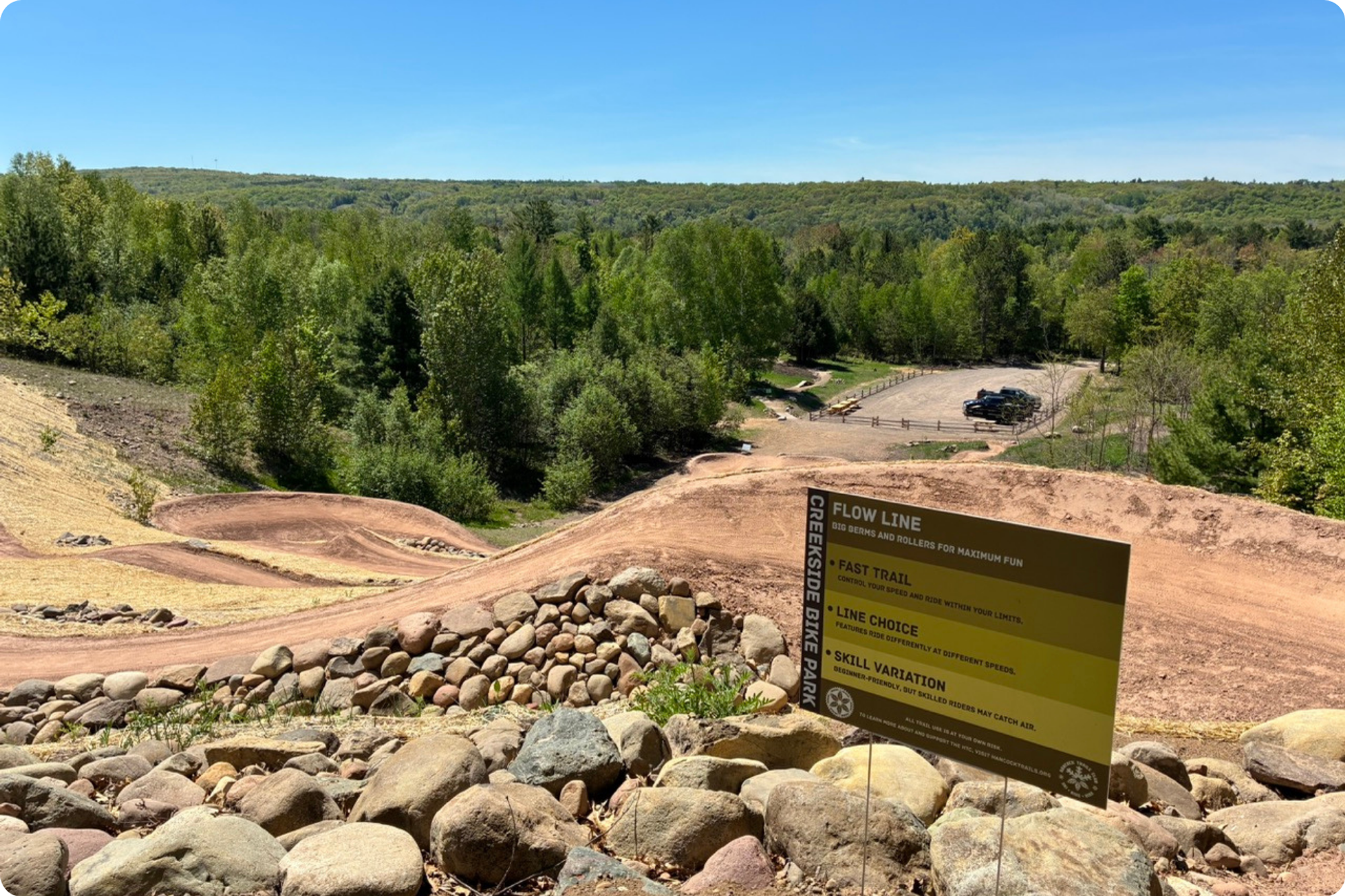 A windy mountain bike trail descending down a hill to a parking lot, with a forest in the background. The foreground includes new Creekside Trailhead and Bike Park signage.