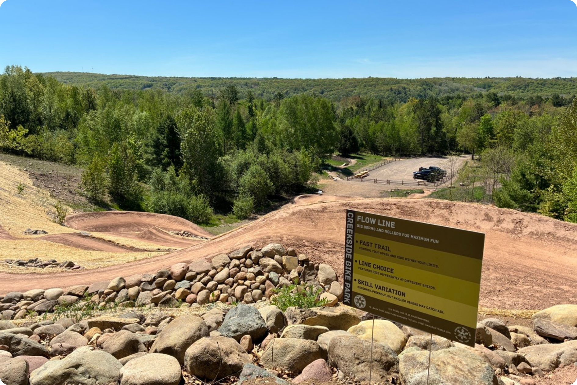 A windy mountain bike trail descending down a hill to a parking lot, with a forest in the background. The foreground includes new Creekside Trailhead and Bike Park signage.