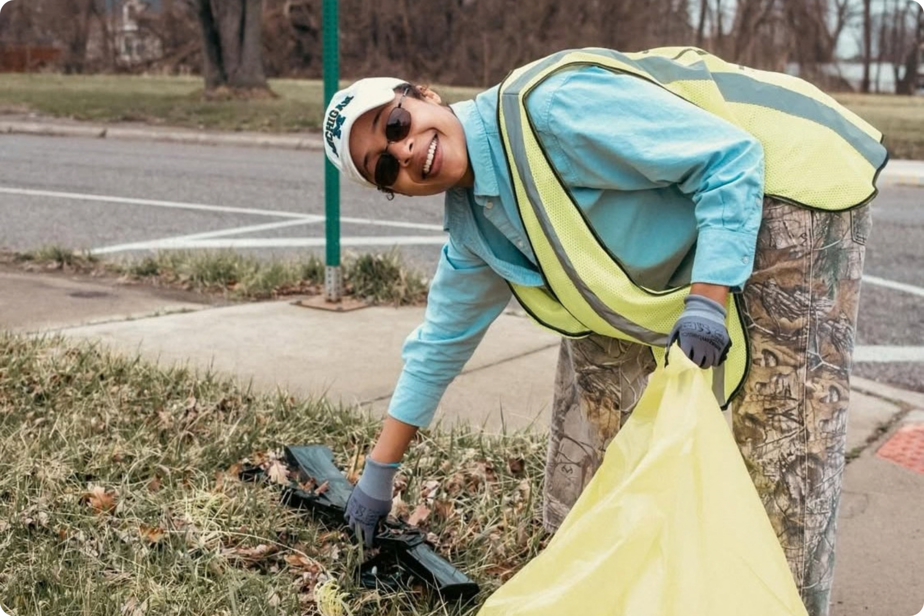 A young woman in a hi-vis vest smiles at the camera while picking up trash from the verge.