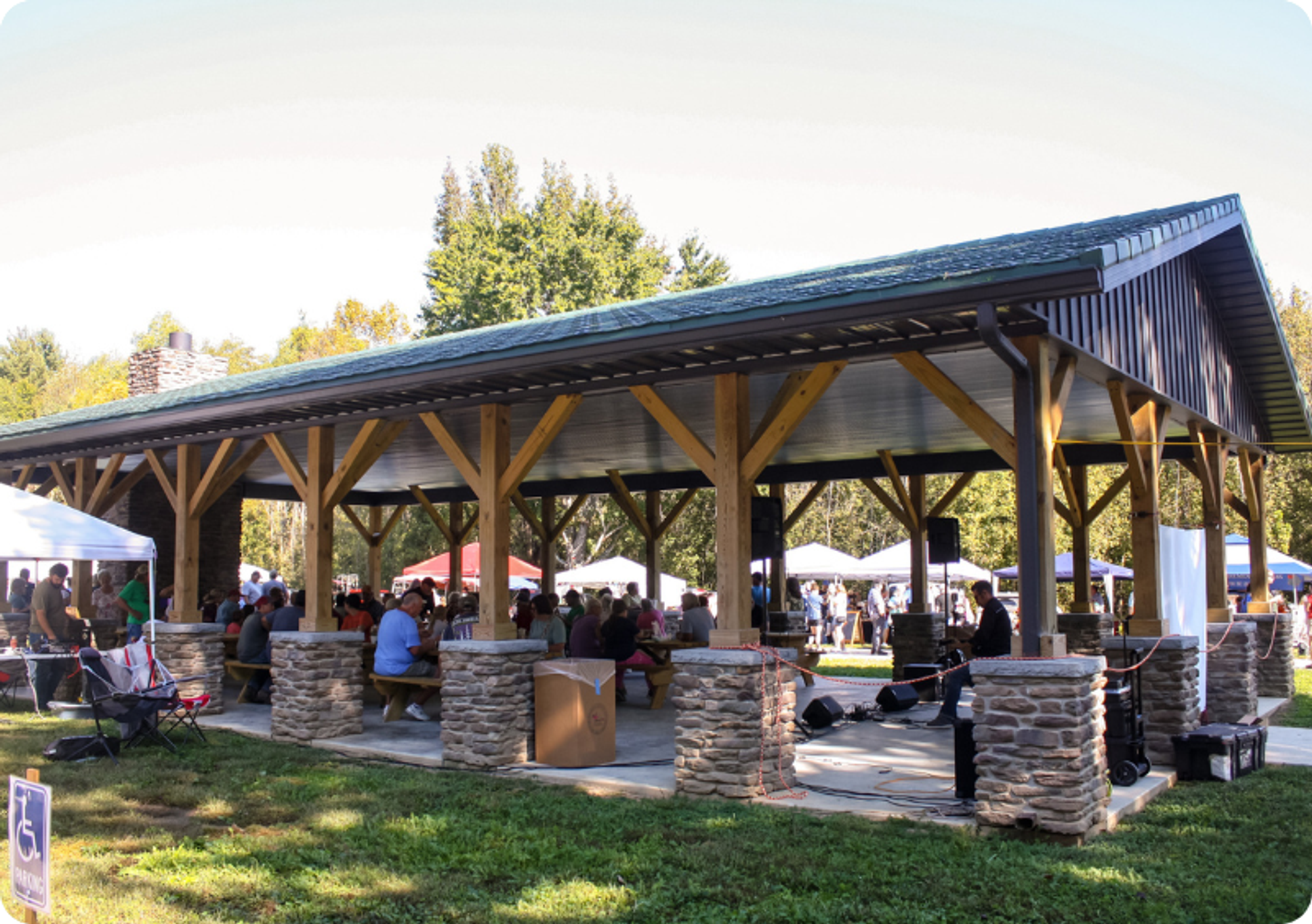 A permanent shelter house in the grass full of people seated at benches, with market gazebos in the background