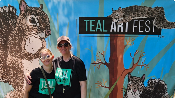 Two Teal Art Fest volunteers stand in front of a banner featuring illustrations of squirrels.