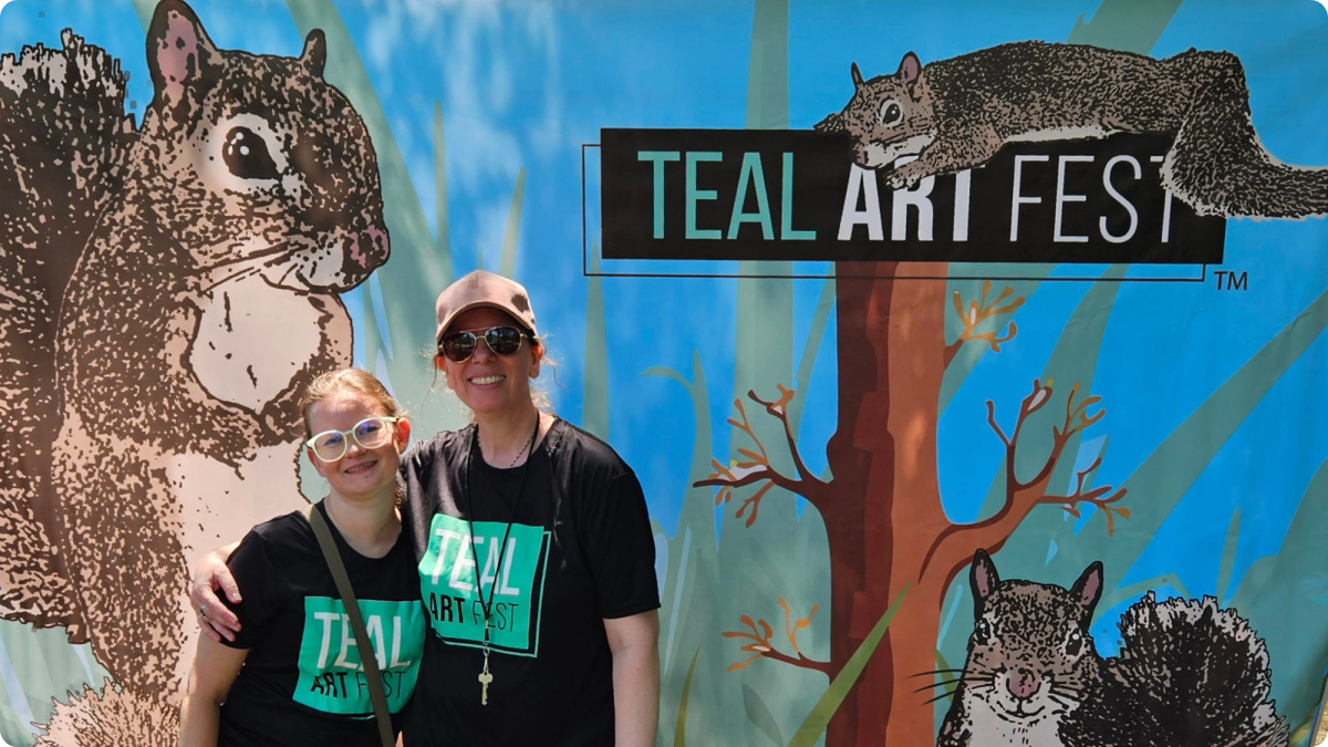 Two Teal Art Fest volunteers stand in front of a banner featuring illustrations of squirrels.