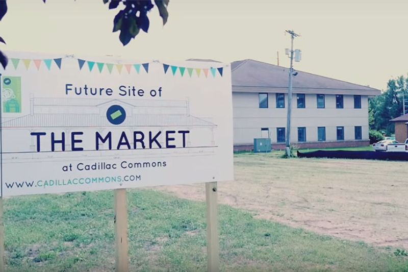 An empty dirt lot stands in the background of a sign that reads, "Future Site of THE MARKET at Cadillac Commons."