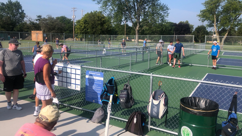 3-4 people play pickleball in each of several pickleball courts. The walkway between the courts is crowded with spectators, athletic equipment, and bags.