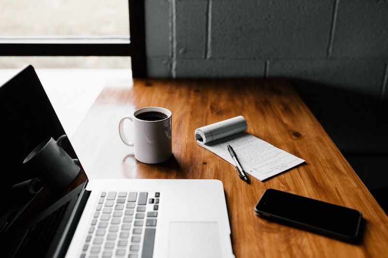 A macbook, a coffee mug, a notepd and an iPhone on a wood table. Photo by Andrew Neel on Unsplash