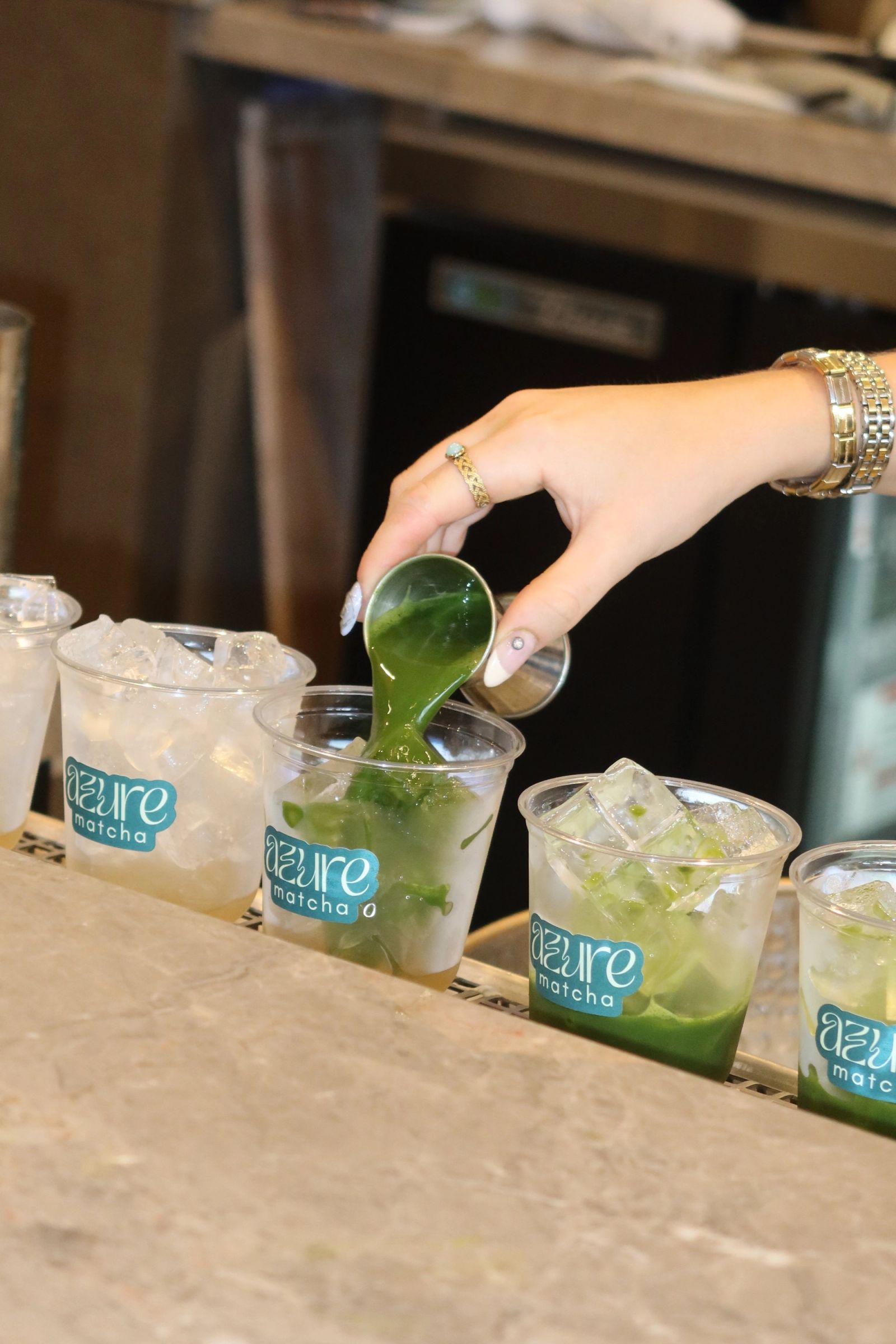 A person pours vibrant green matcha liquid from a pitcher into a glass filled with ice and matcha at an Azure Matcha establis