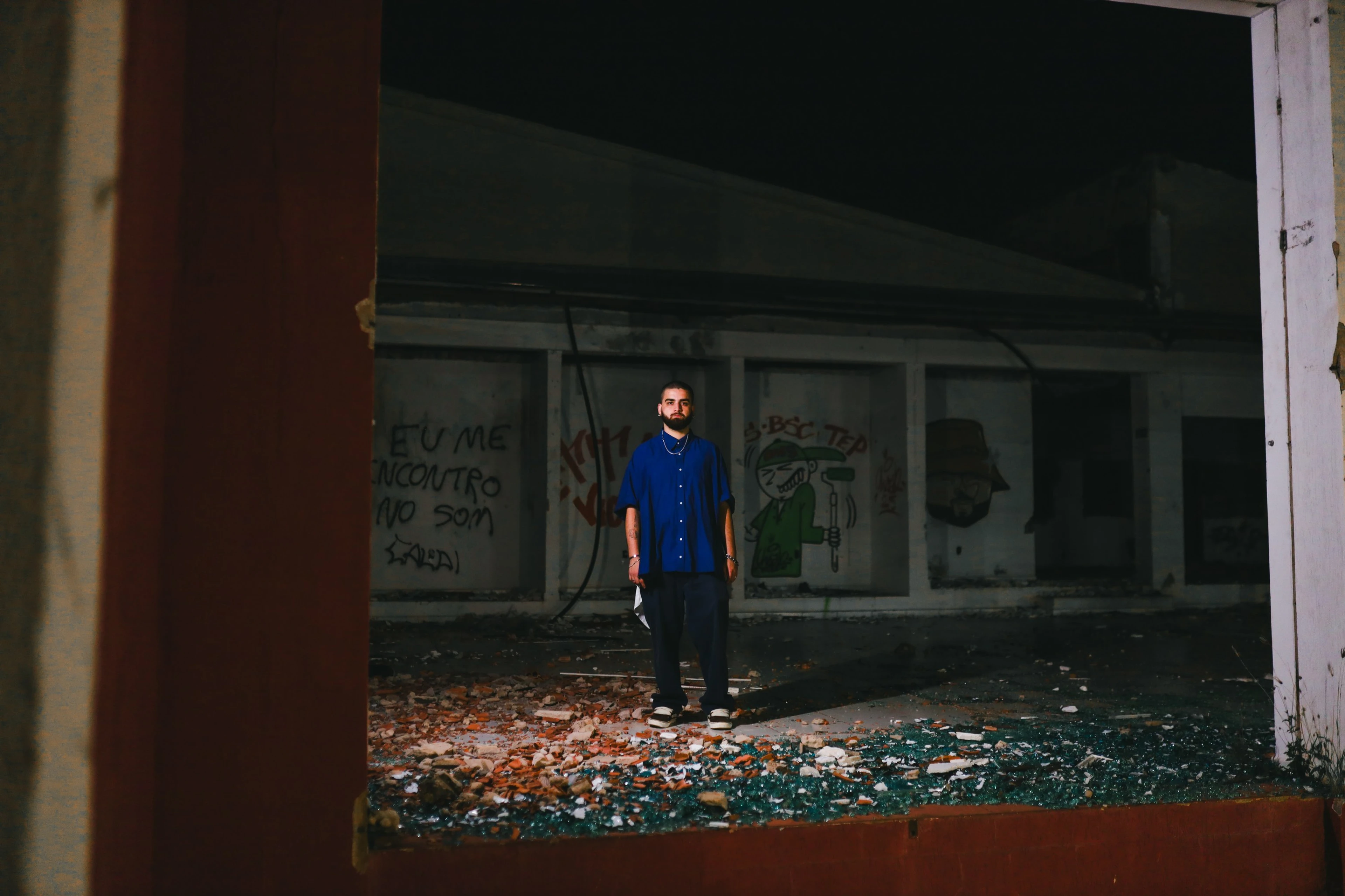 Homem de camisa azul de pé dentro de uma construção abandonada, à noite