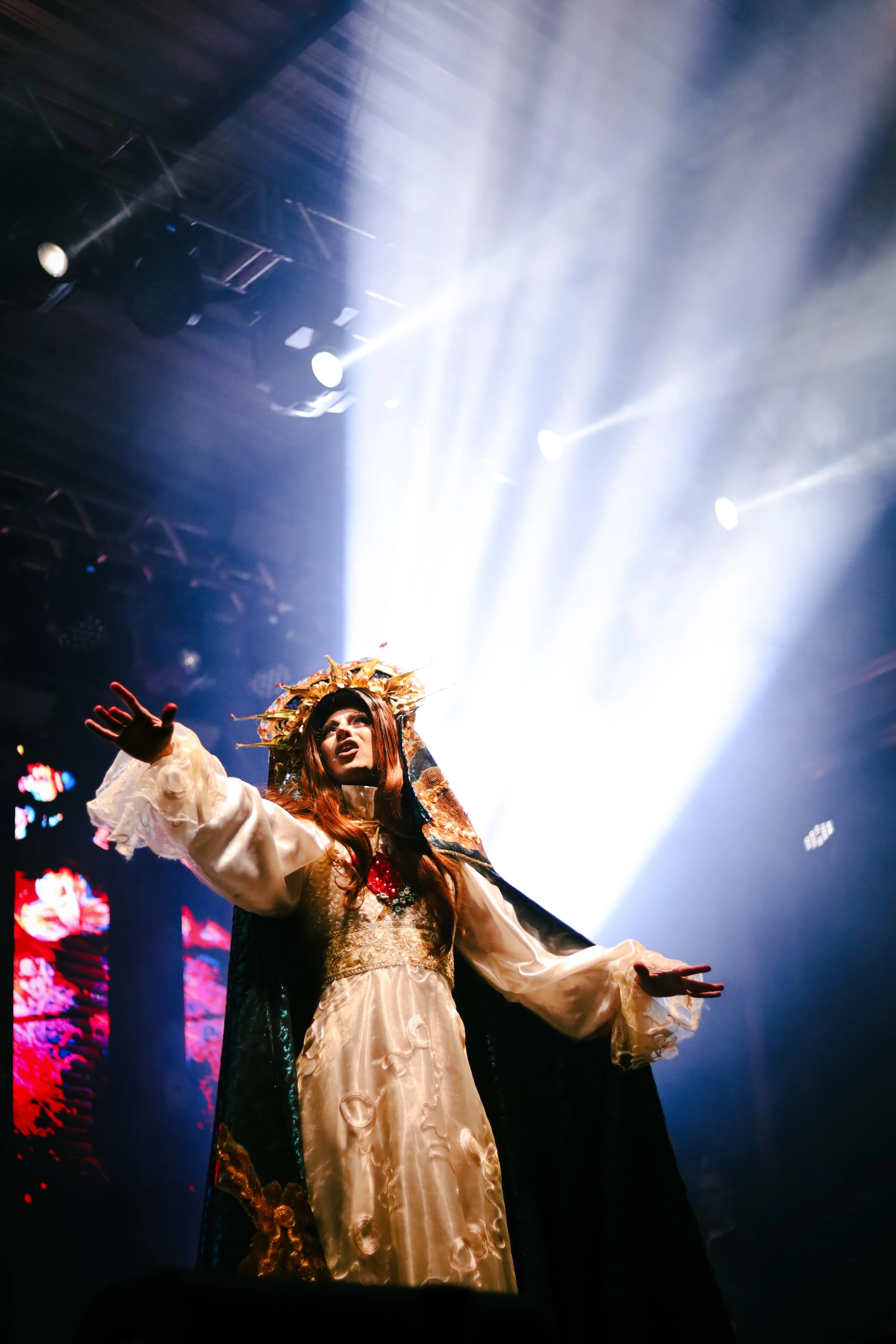 Artista de vestido de renda e coroa de flores no palco, com feixes de luz branca ao fundo