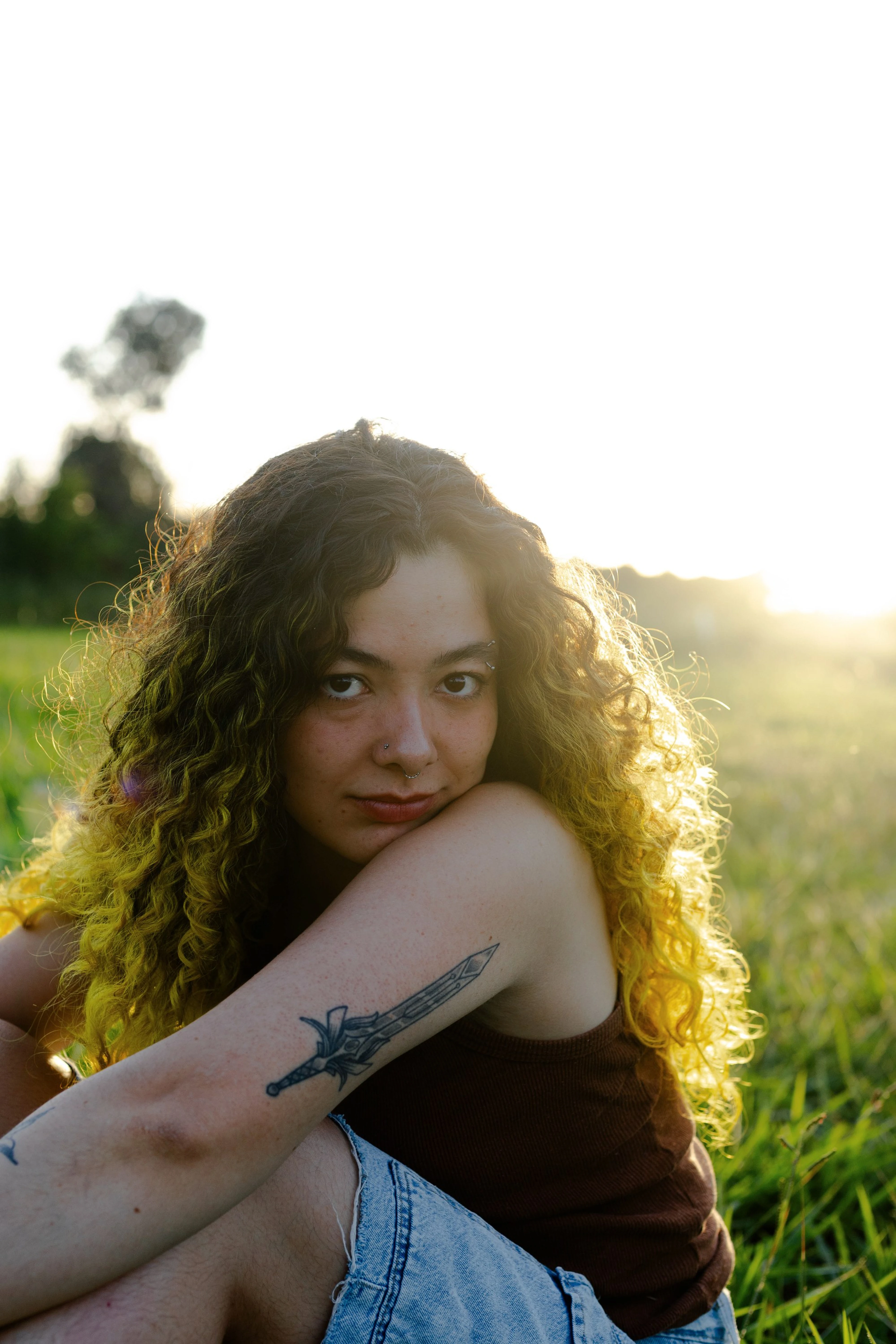 Mulher de cabelo cacheado sentada na grama, com o sol por trás
