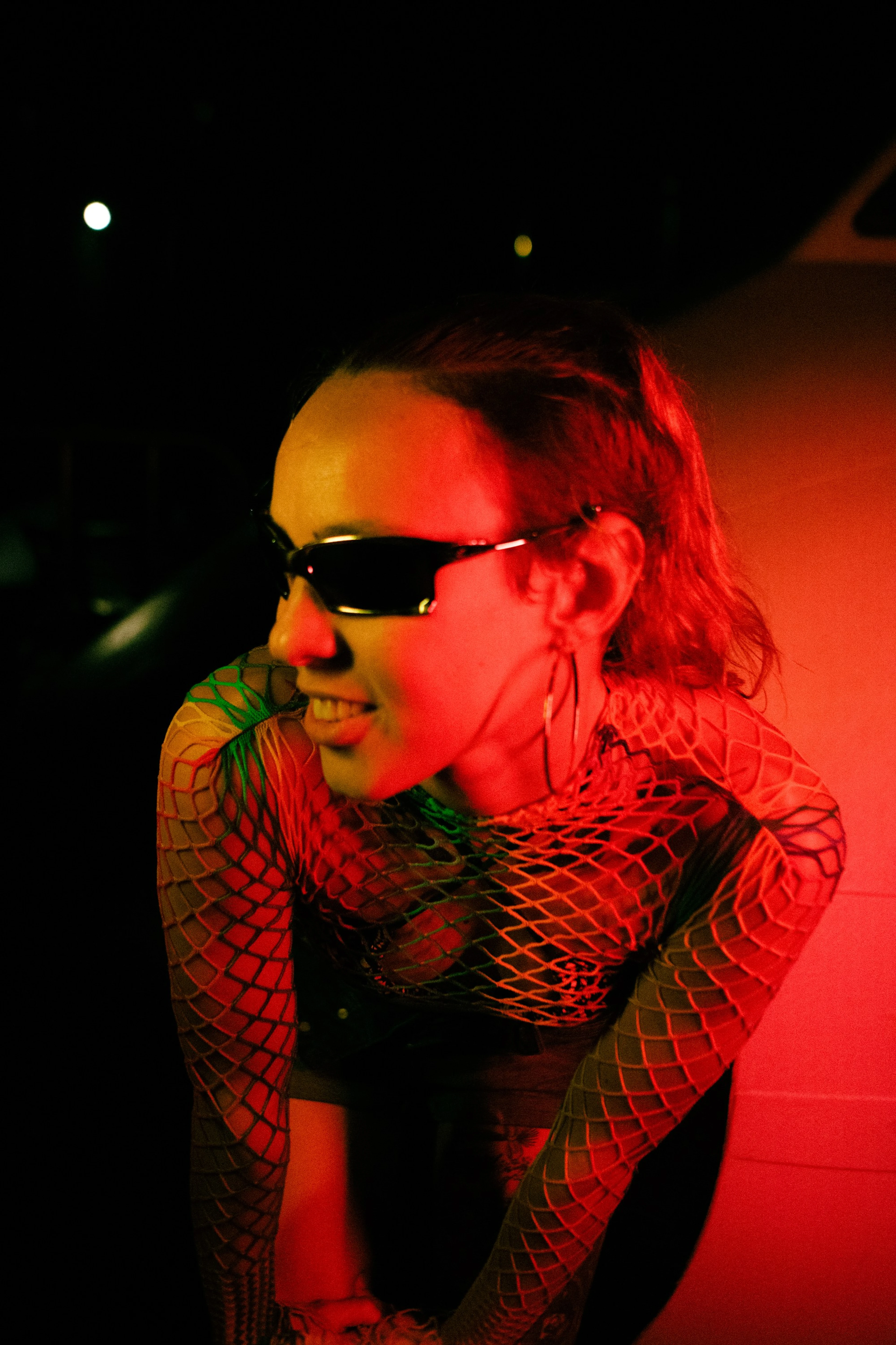 Mulher de óculos escuros e blusa de rede sorrindo sob luz vermelha