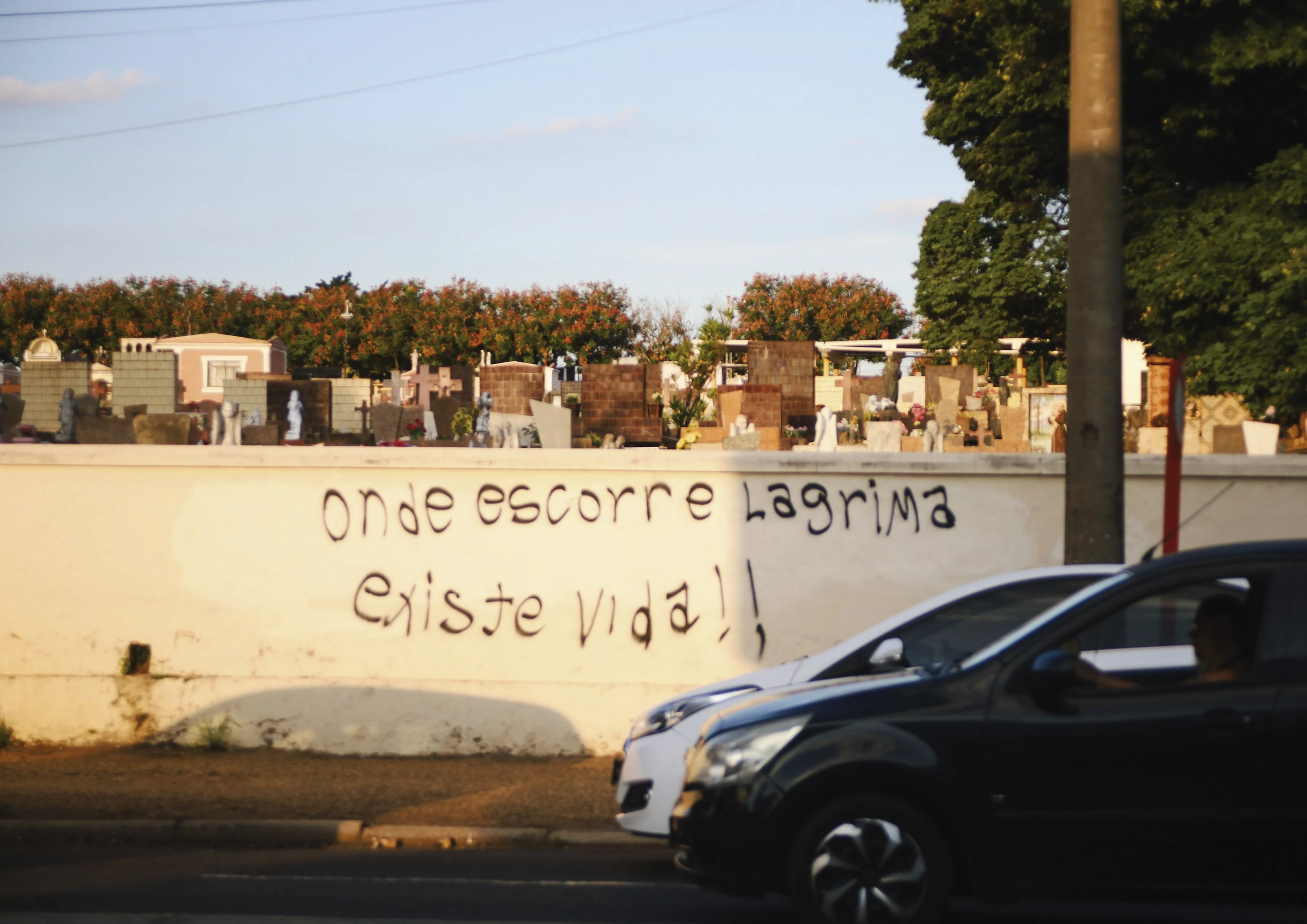 Muro de cemitério com a frase “Onde escorre lágrima existe vida!” e um carro passando