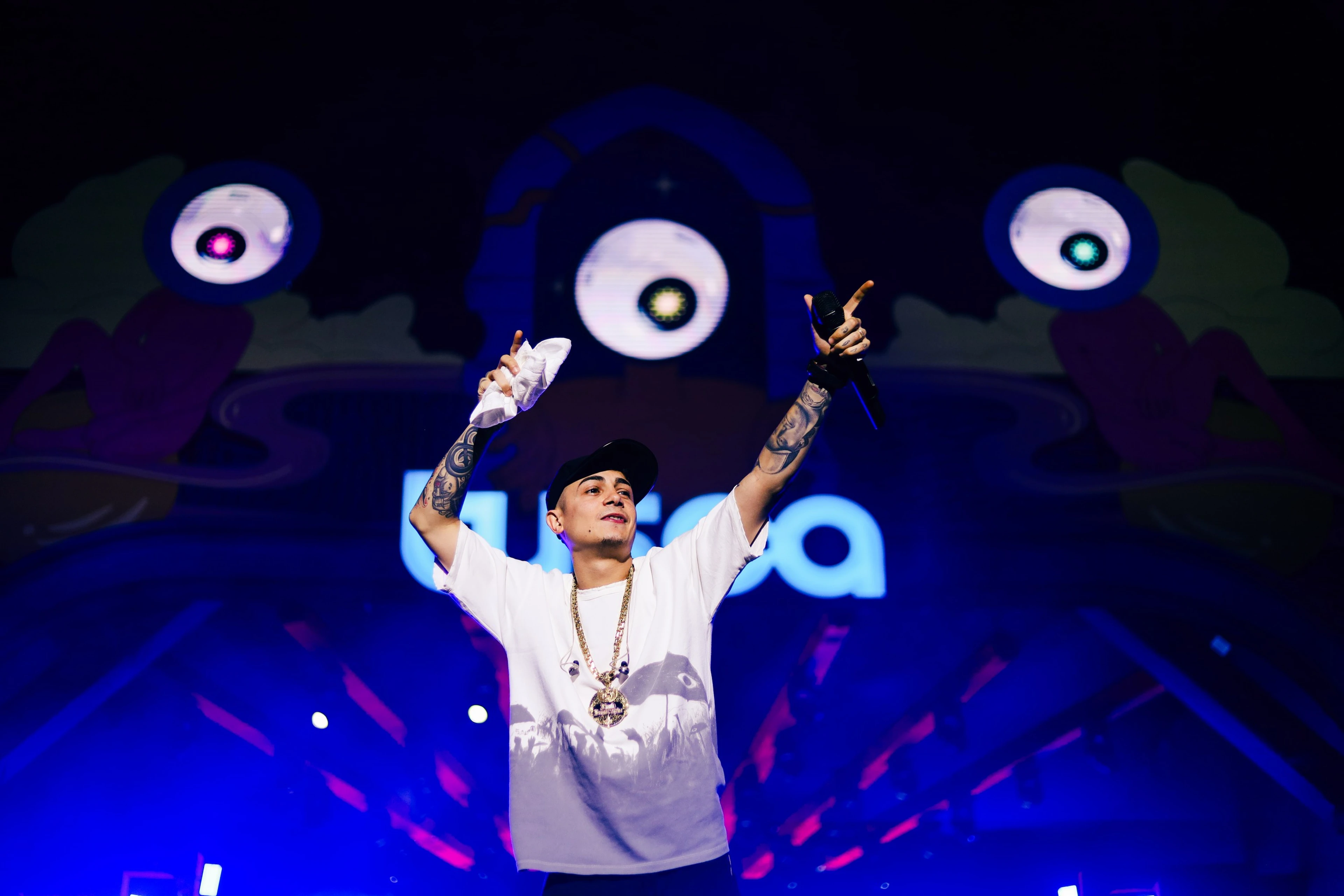 Artista de boné e camiseta branca com os braços erguidos no palco, luz azul ao fundo