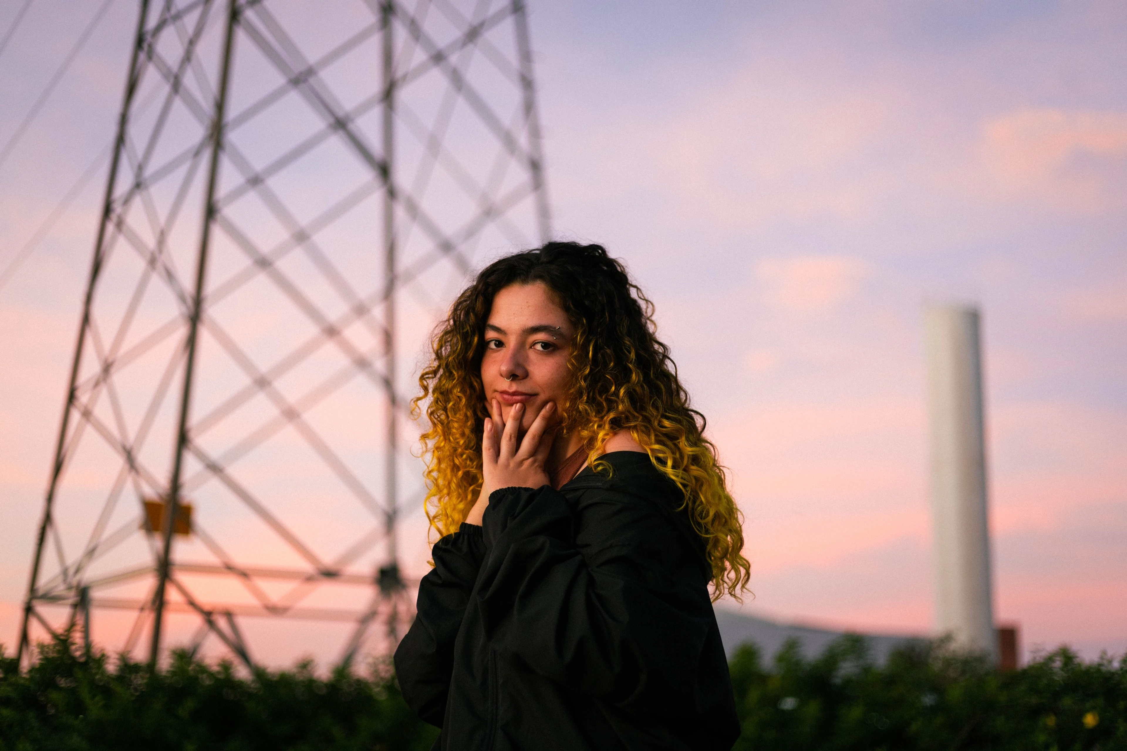 Retrato de mulher de cabelo cacheado ao pôr do sol, com uma torre de energia ao fundo