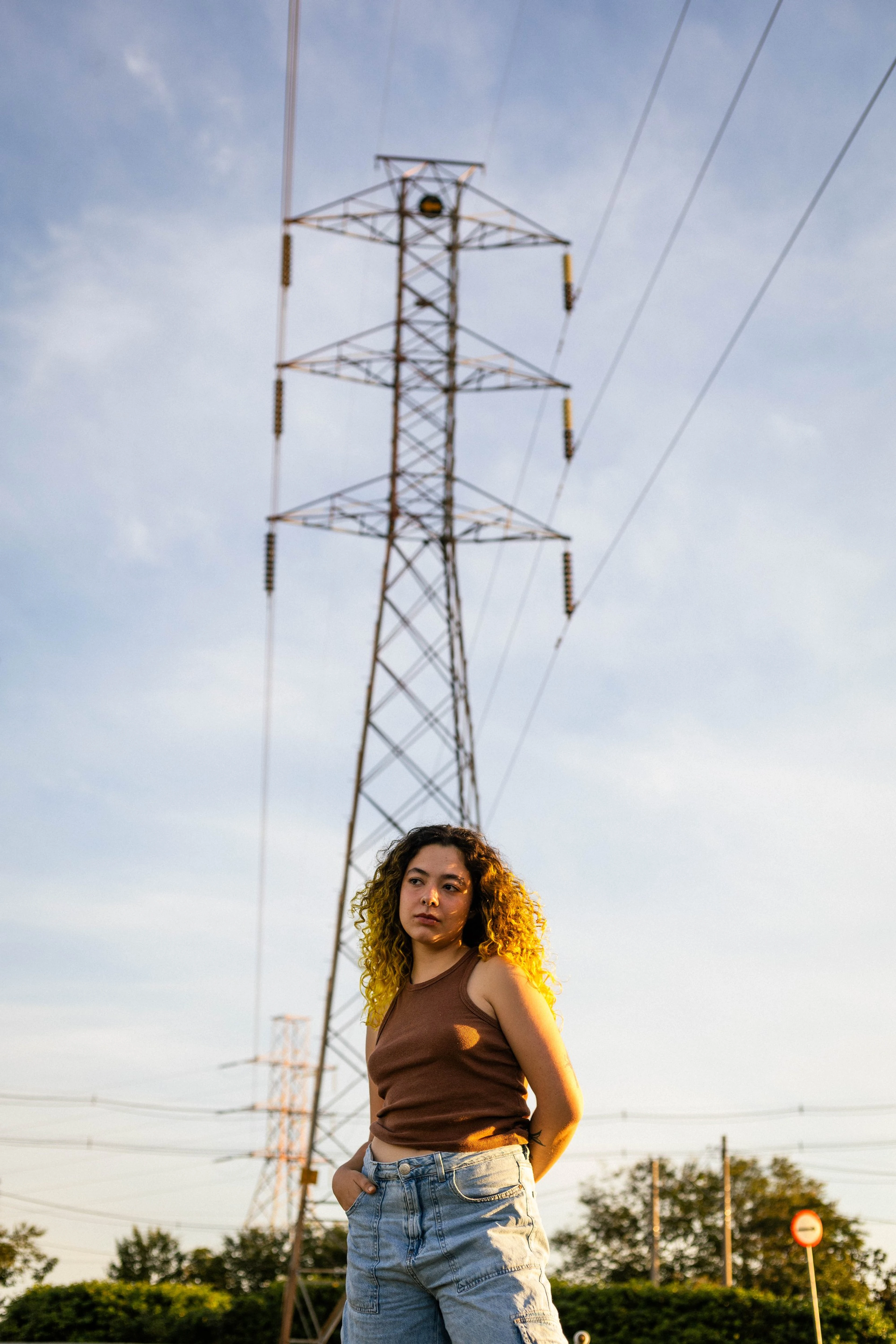 Mulher de cabelo cacheado e regata marrom diante de uma torre de energia, céu azul