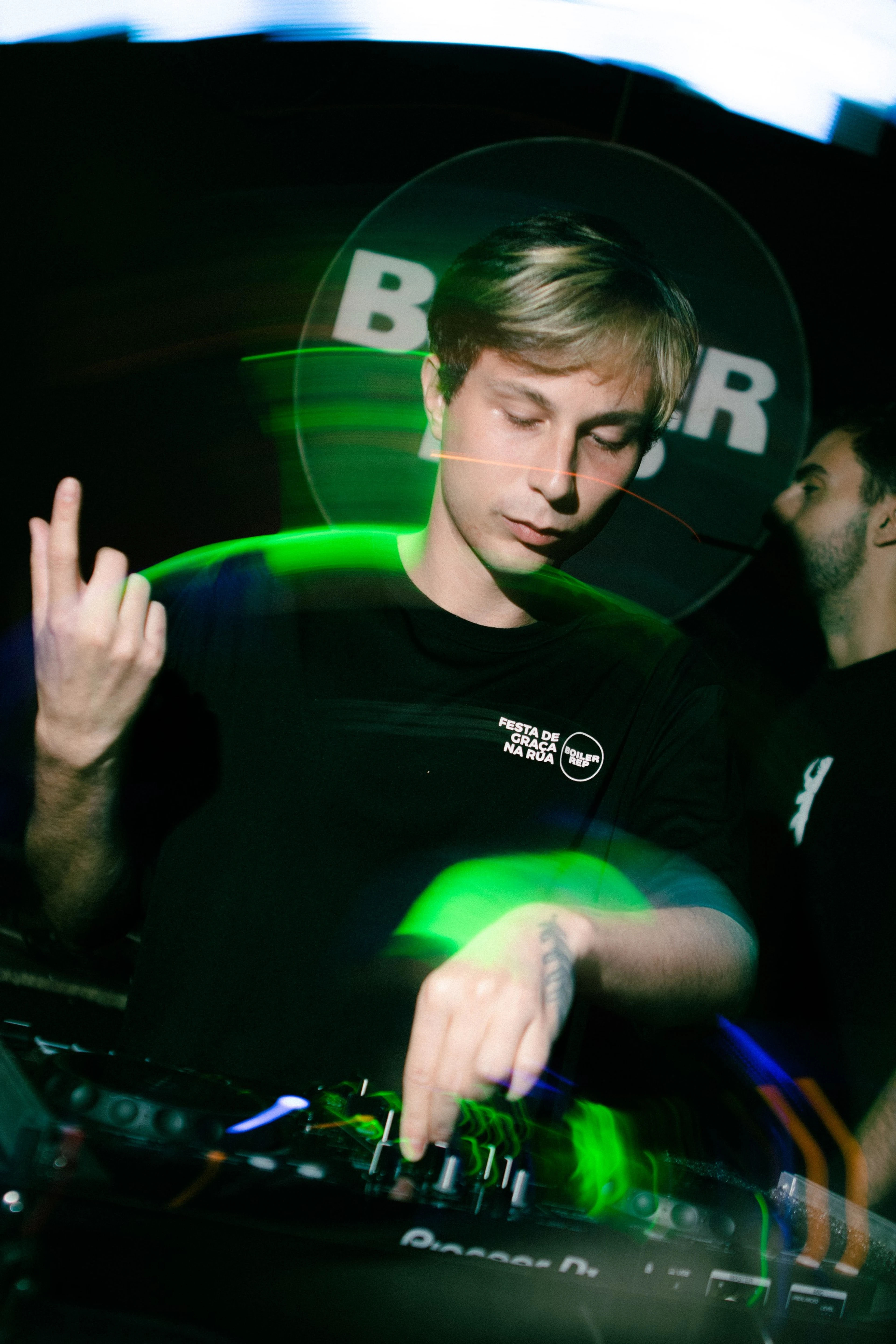 DJ loiro de olhos fechados tocando, com rastros de luz verde
