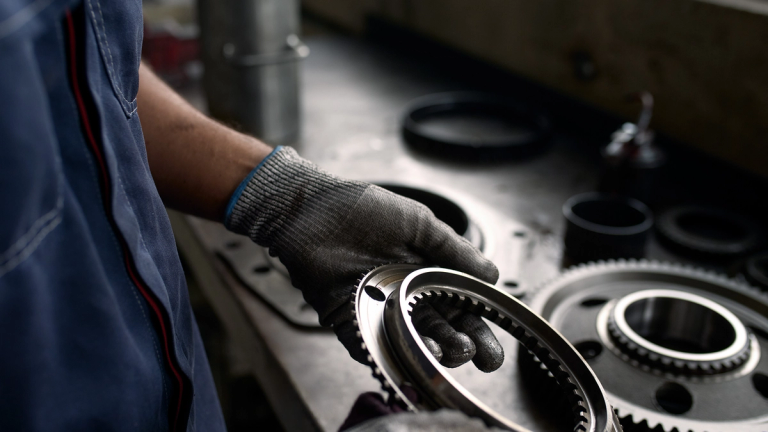 Worker wearing protective gloves holding a metal gear component, with other mechanical parts and tools laid out in the background.