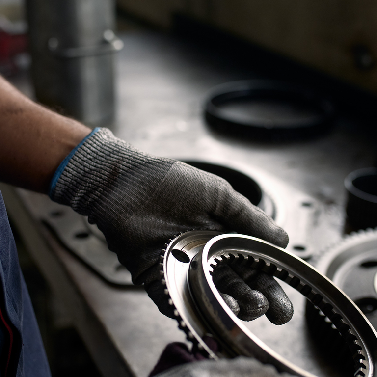 Worker wearing protective gloves holding a metal gear component, with other mechanical parts and tools laid out in the background.
