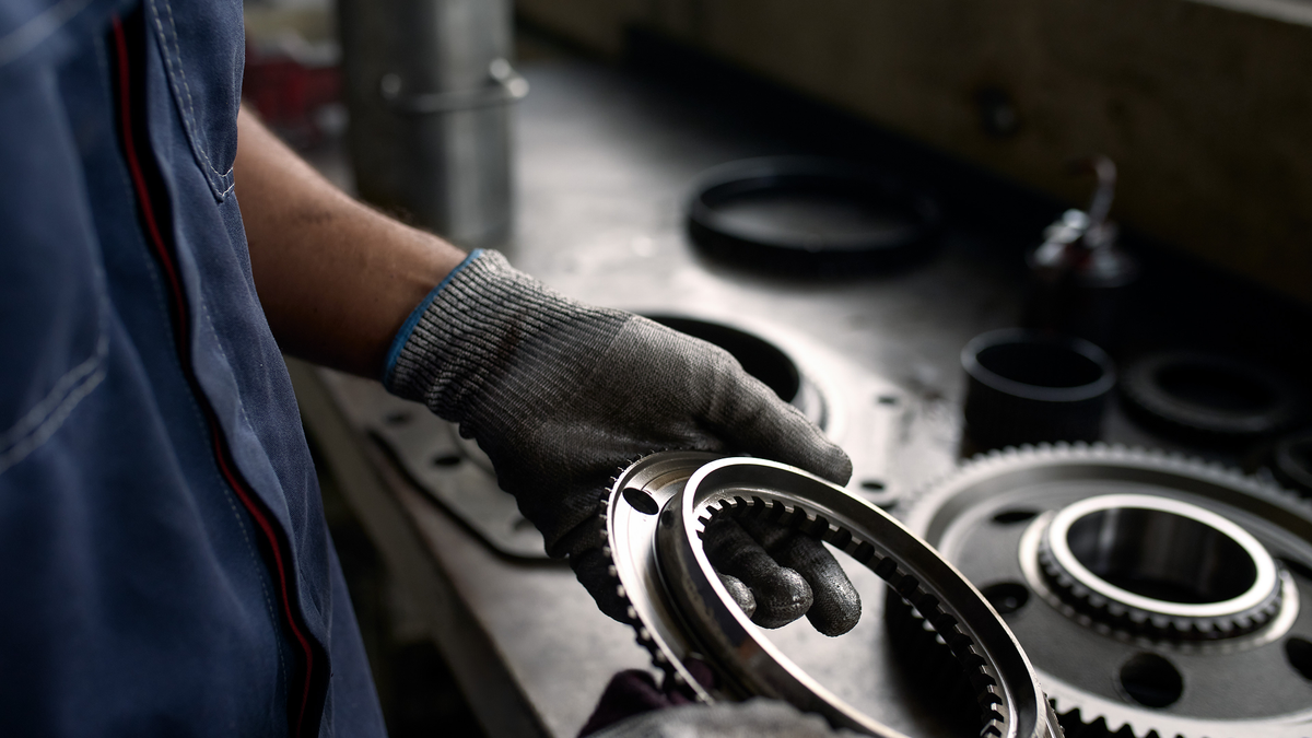 Worker wearing protective gloves holding a metal gear component, with other mechanical parts and tools laid out in the background.