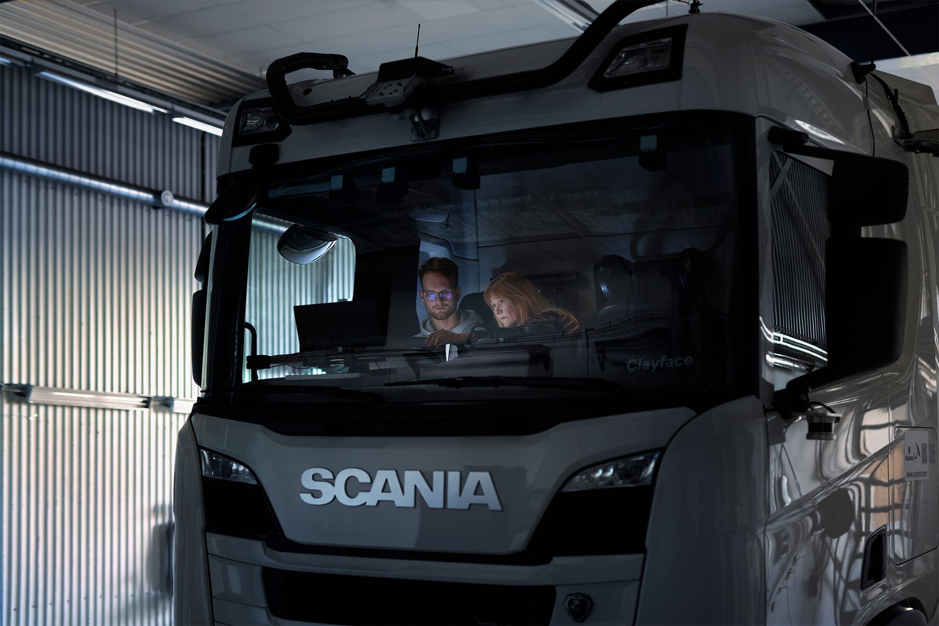 Two people sitting in the cab of a white Scania truck viewed through the windshield in an industrial warehouse setting.