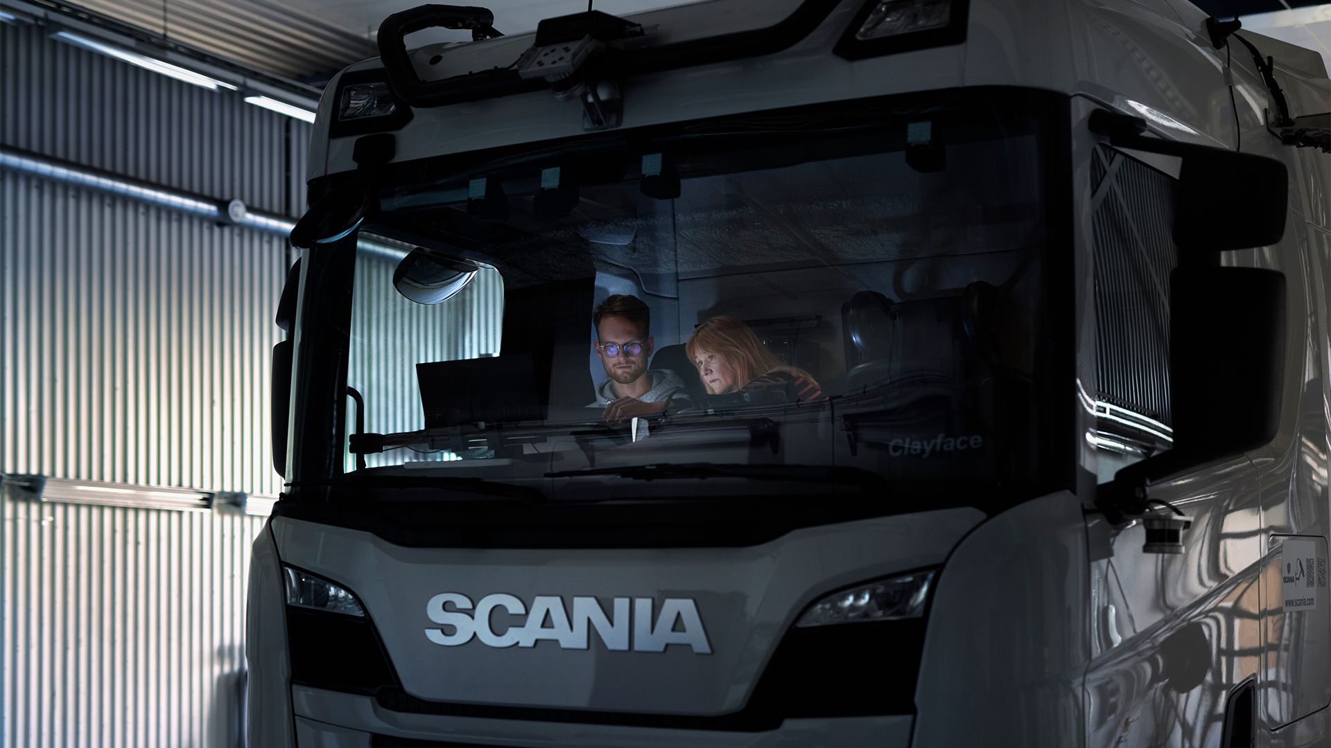 Two people sitting in the cab of a white Scania truck viewed through the windshield in an industrial warehouse setting.
